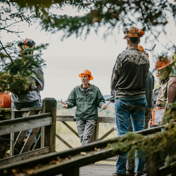 Forestry professor in raincoat on a deck next to a lake, talking to a group of students, in raincoats and hard hats
